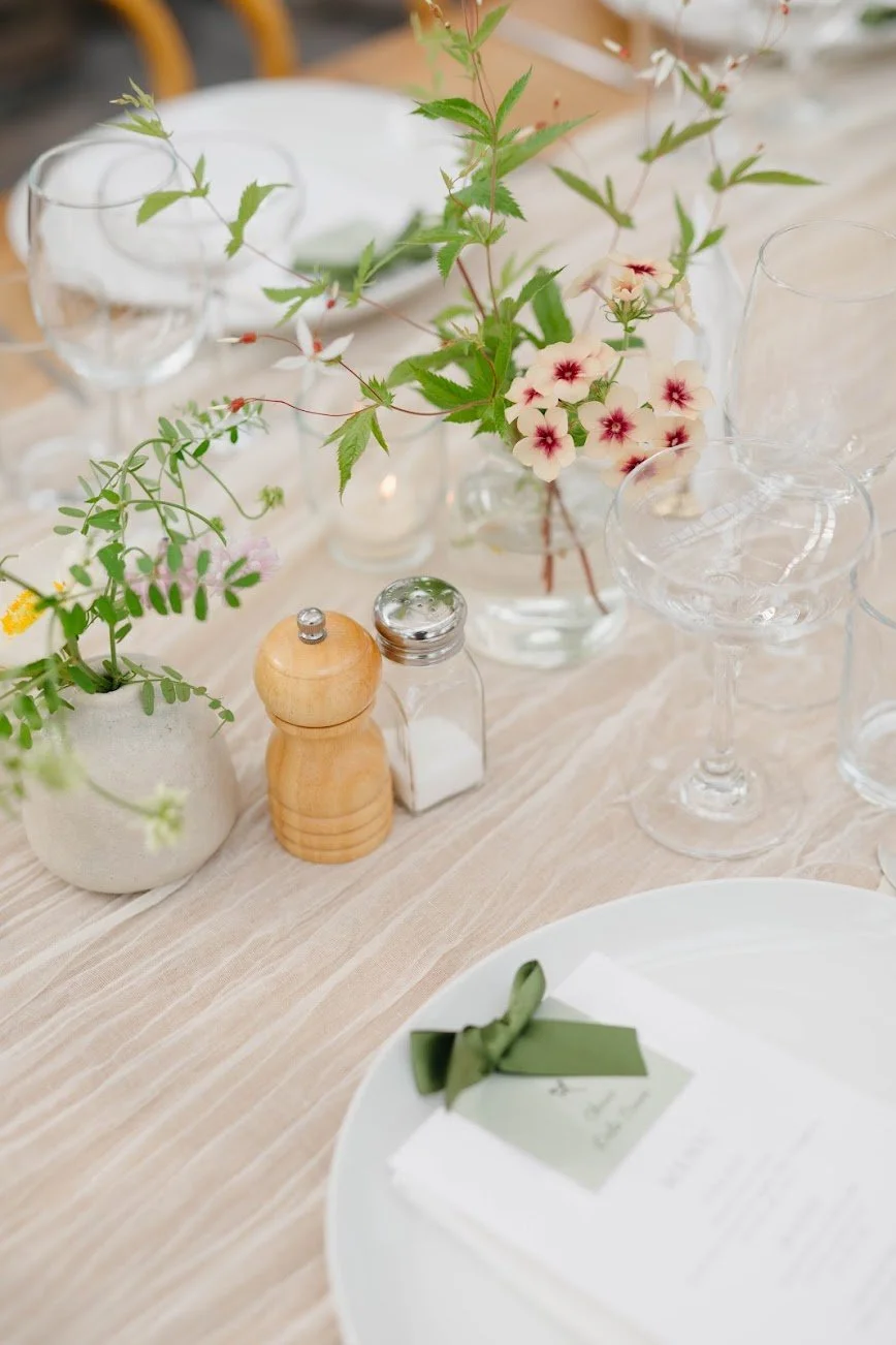 Delicate purple blossoms sit in glass bud vase with petite round leaves on fabric draped table