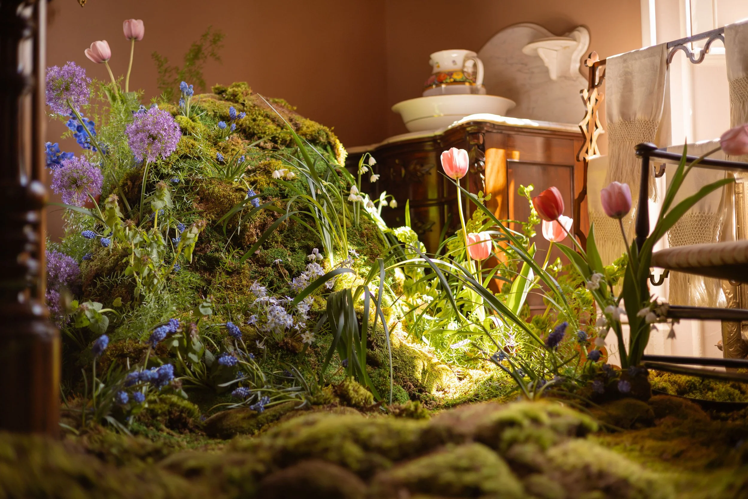 Lump of moss with vibrant tulips, allium, forget-me-nots and more local blooms, beside wooden furniture.