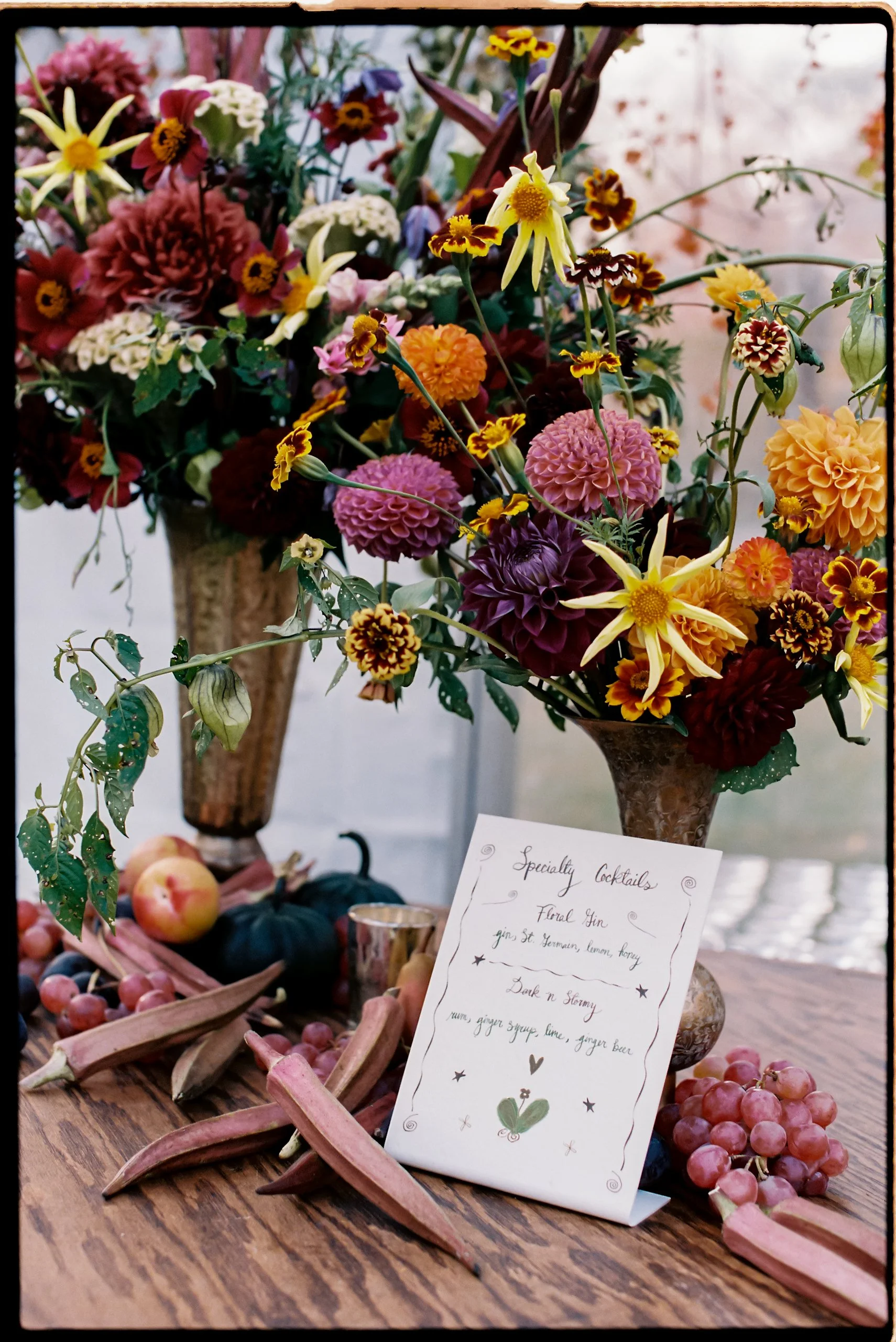 A bouquet of colorful flowers in a tall vase and a handwritten menu card on a wooden table surrounded by grapes, apples, and breadsticks.
