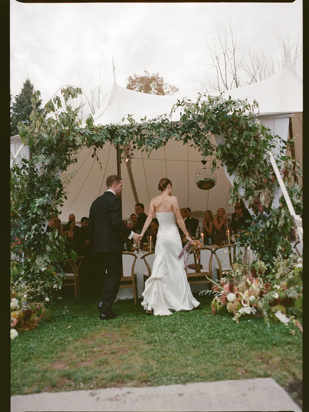 Couple walking beneath a vine-covered tent entrance filled with organic greenery and soft seasonal blooms.