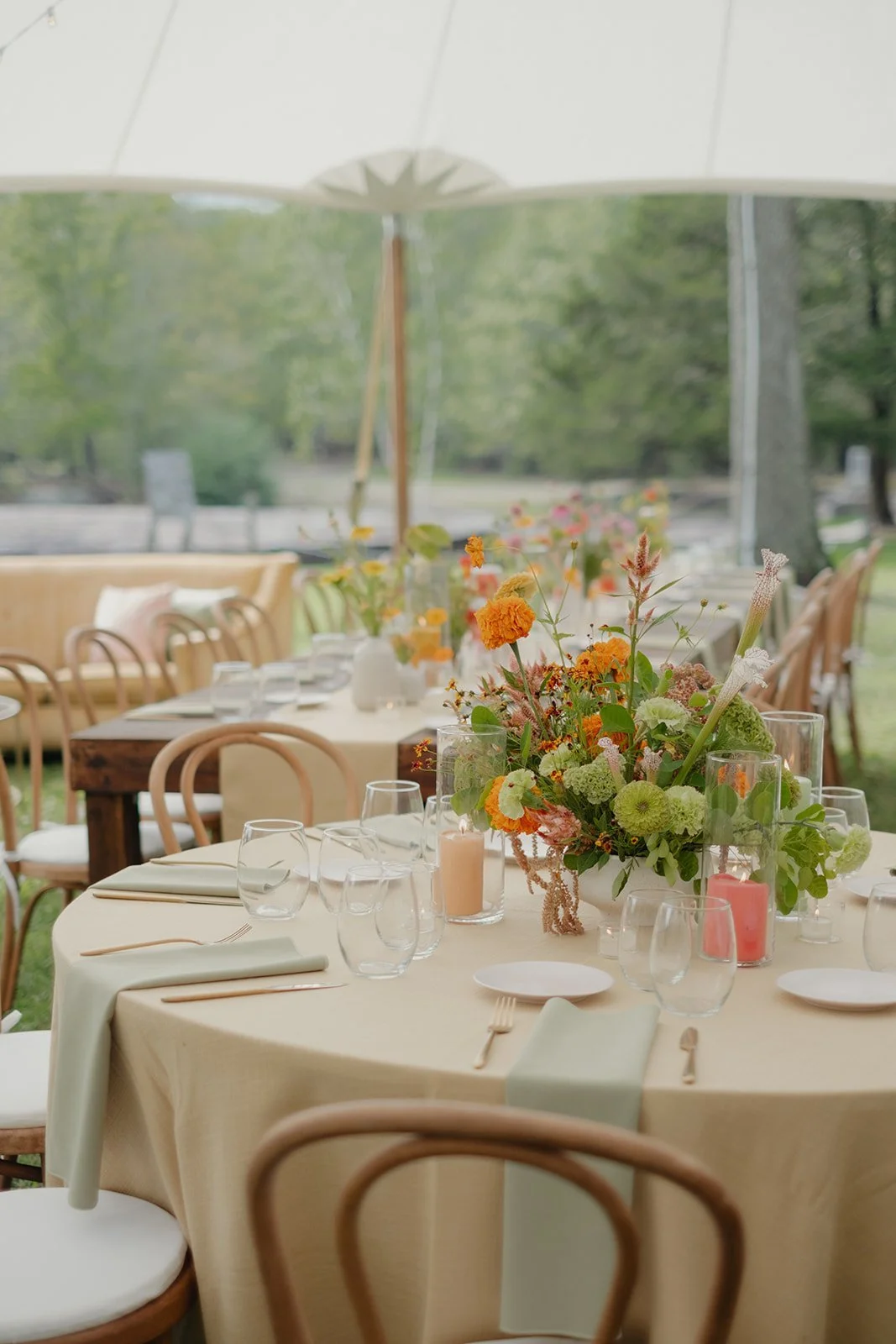 Elegant outdoor event setup with round tables decorated with floral centerpieces, glassware, candles, and napkins under a large white umbrella in a lush garden setting.