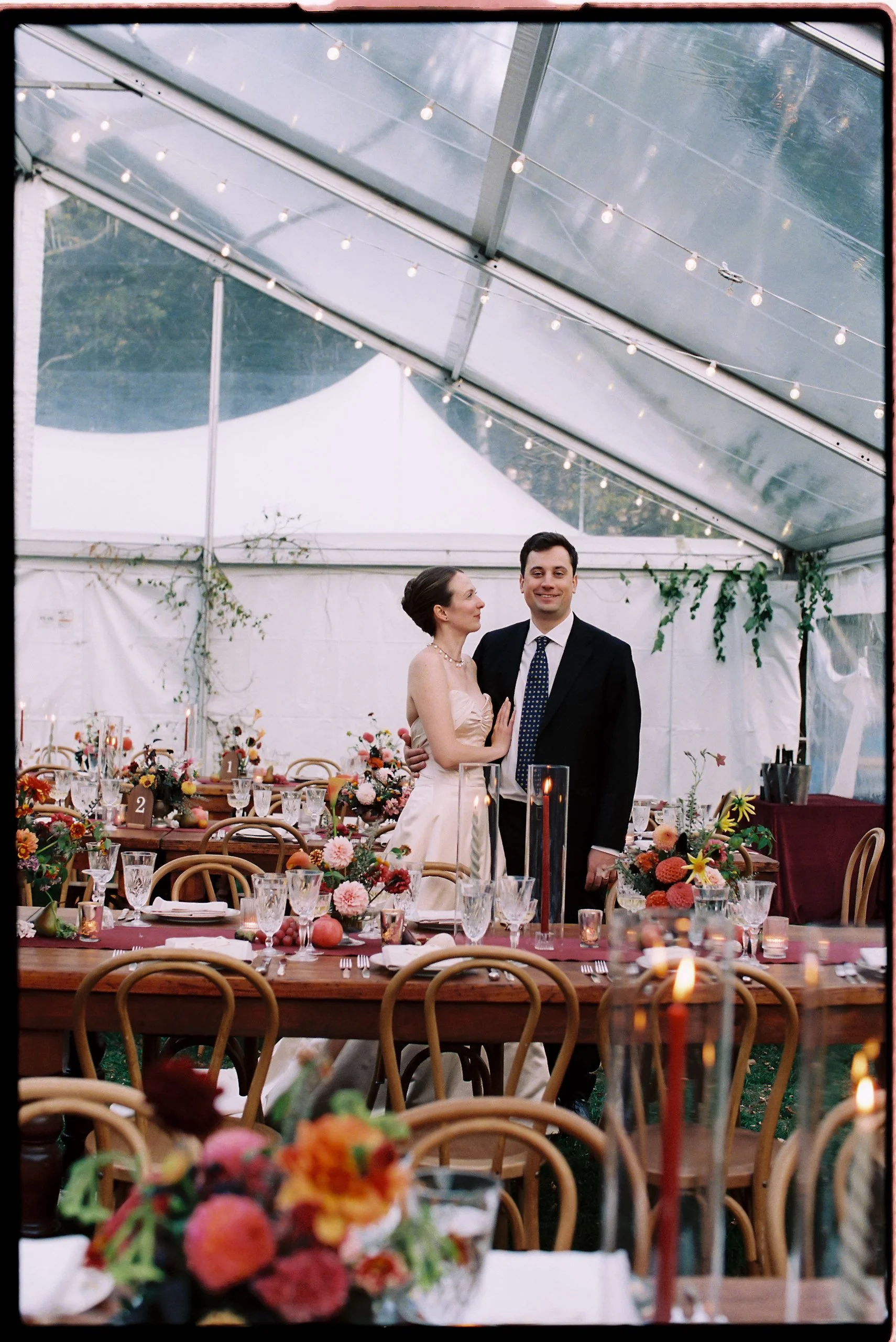 Couple standing among long reception tables adorned with lush fall floral centerpieces beneath a clear tent