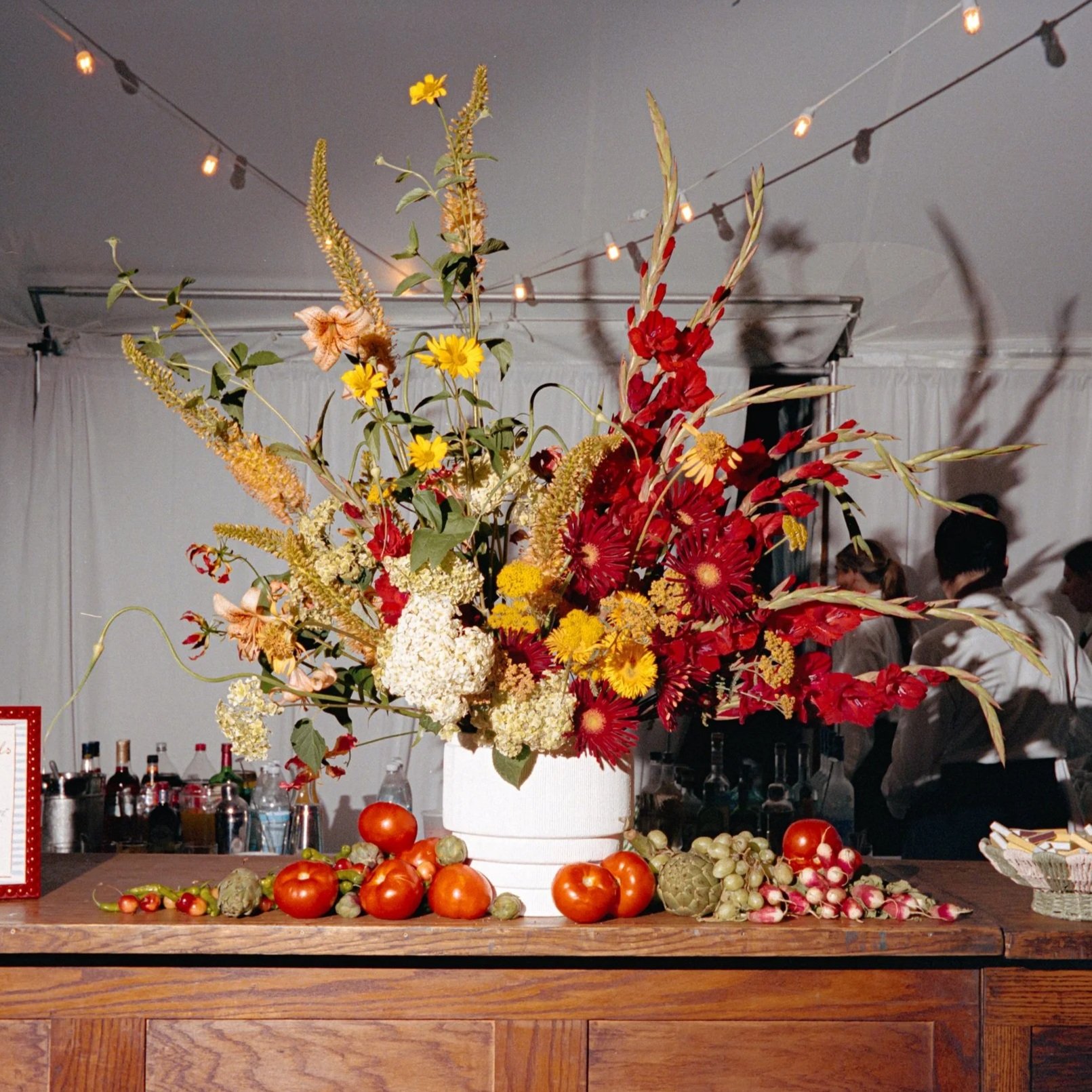 A large floral arrangement with red, yellow, and white flowers in a white vase, surrounded by tomatoes, grapes, artichokes, radishes, and other produce on a wooden table in front of a white draped wall.