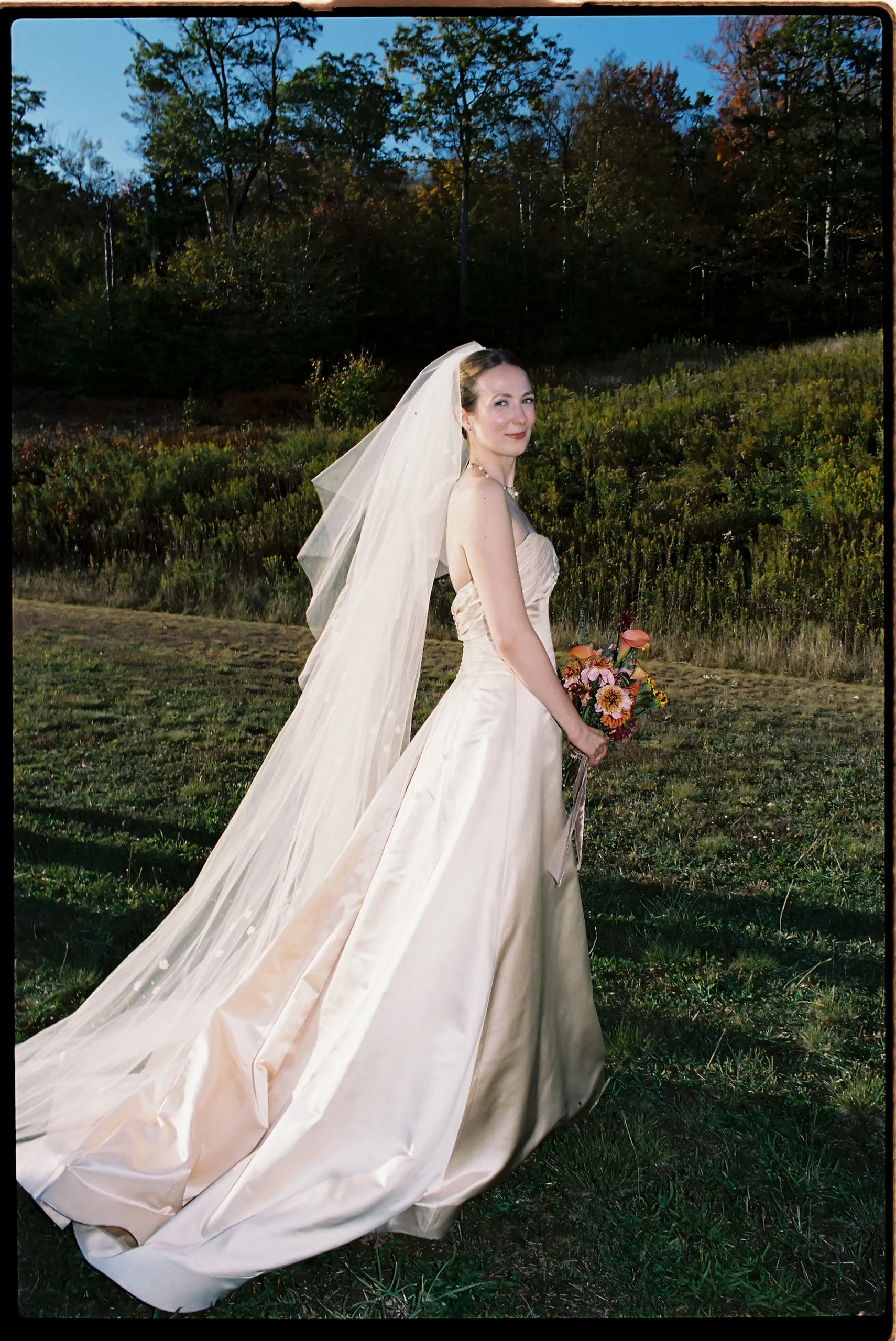 Bride in an open meadow holding a warm-toned floral bouquet with dahlias and soft ribbon accents