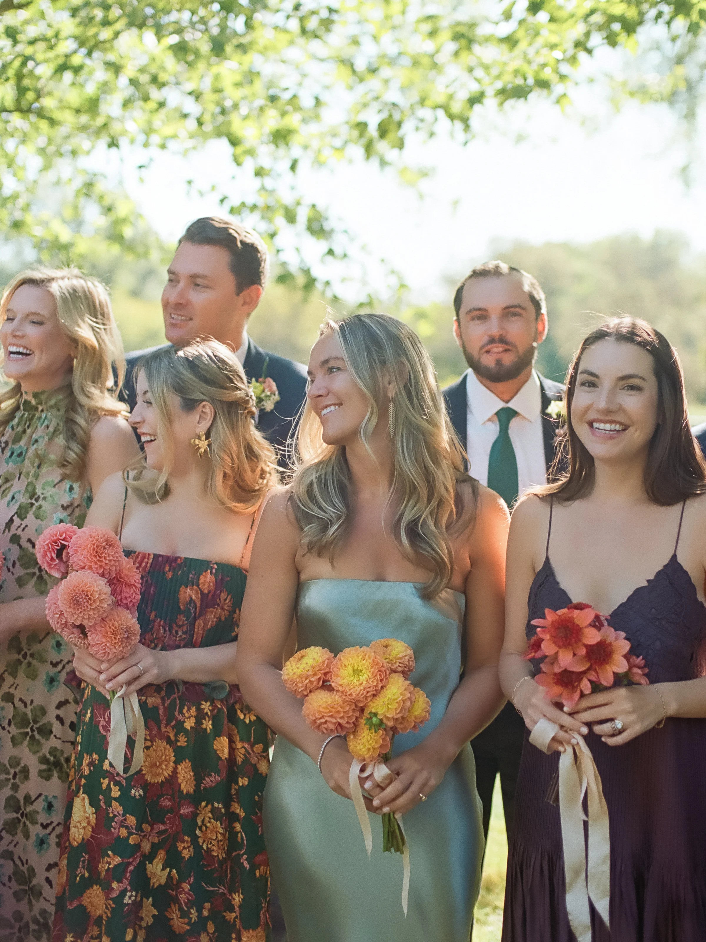 Bridesmaids and groomsmen smiling together beneath trees holding colorful bouquets