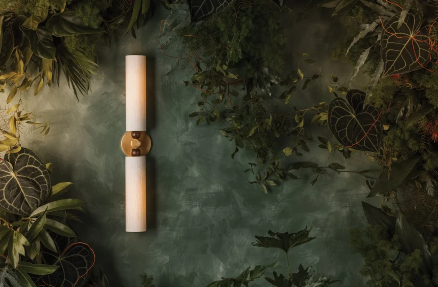 A modern wall-mounted light fixture with a horizontal cylindrical shape, placed on a green textured wall surrounded by various green and dark leafy plants.