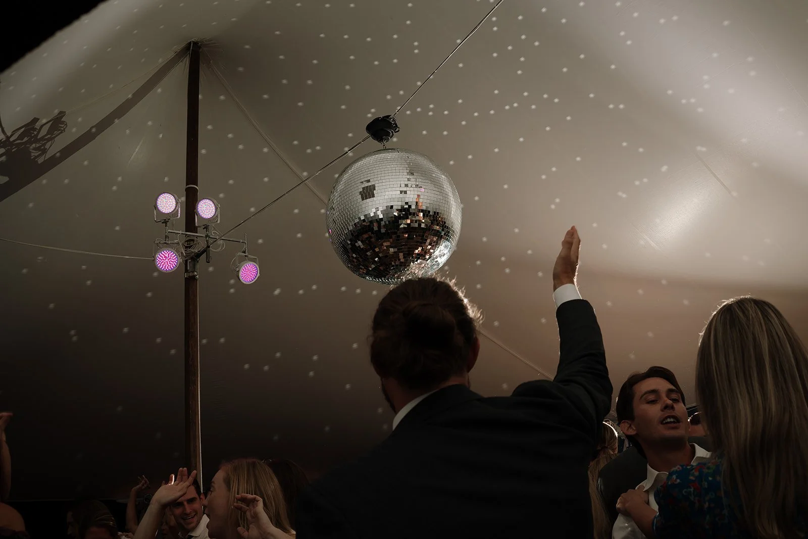 Wedding guests dancing beneath a tent with soft floral accents and shimmering disco ball lighting during the evening reception