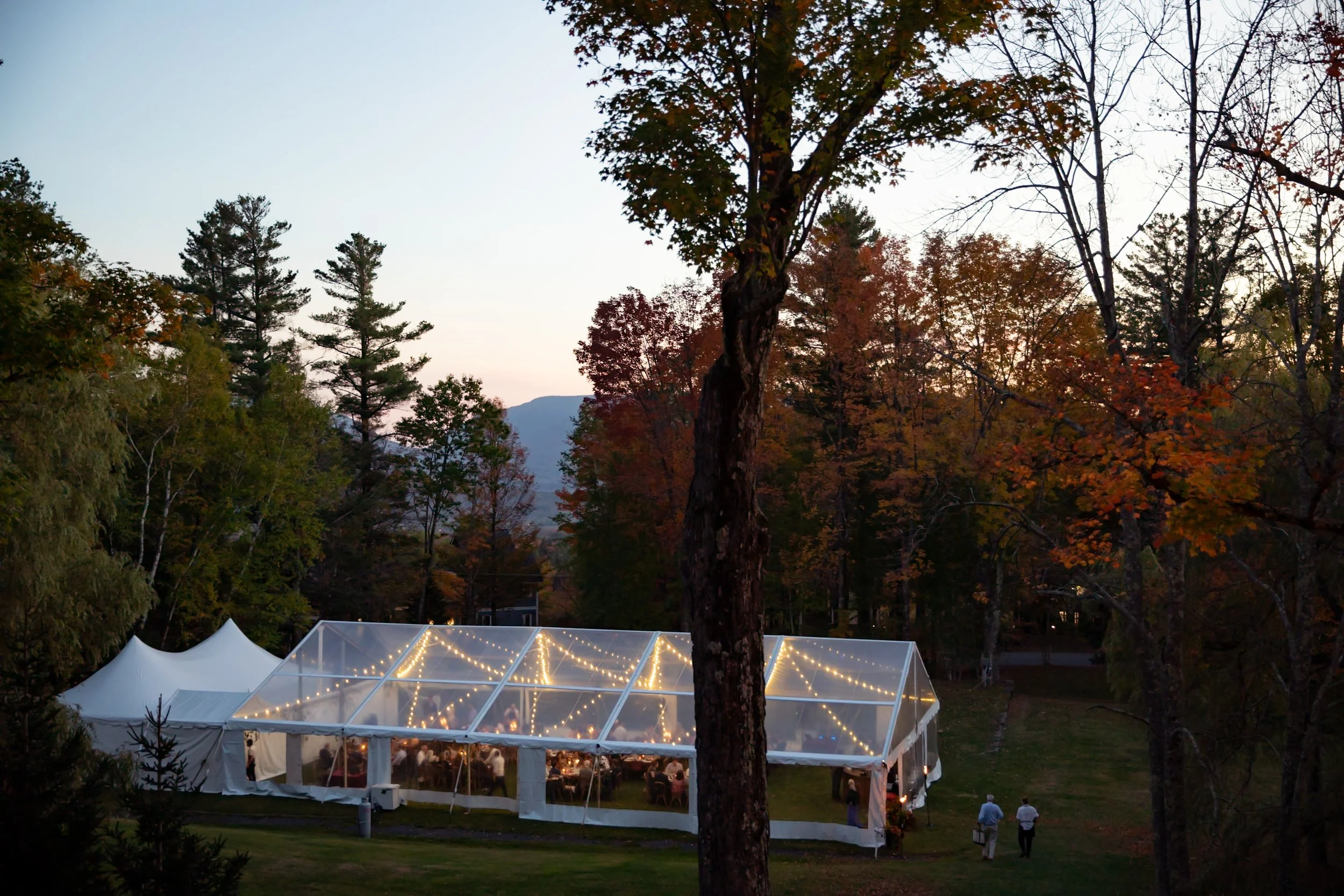 Clear-top reception tent glowing at dusk, framed by autumn trees and softly lit floral-filled tables inside