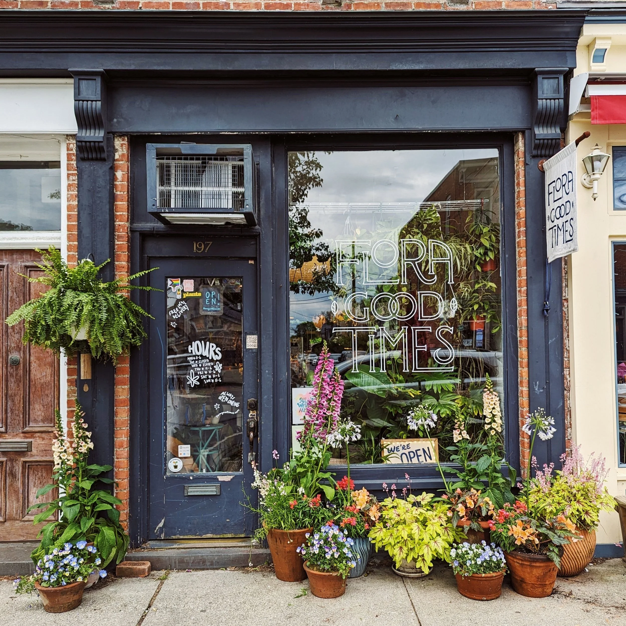 A shop on Beacon, NY's main street with an aged black facade displays FLORA GOOD TIMES on the window. Various plants and flowers are arranged in pots outside. A sign indicates the shop is open. A wooden door is to the left of the entrance.
