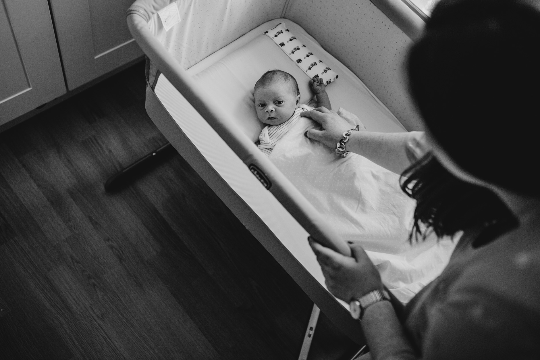 Black & White photo of newborn baby in his cot with, his mother looking at him and touching him with her hand