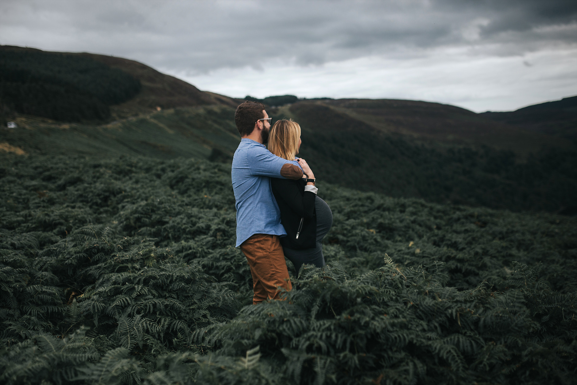 Husband hugging his pregnant wife while looking at the beautiful green irish landscape