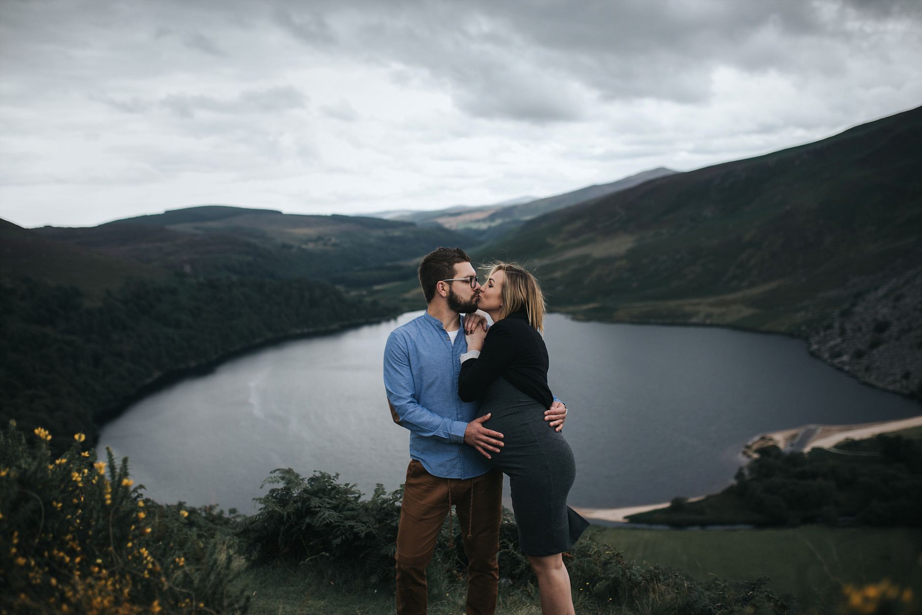Couple kissing in front of the lake while husband touches his wife’s pregnant belly