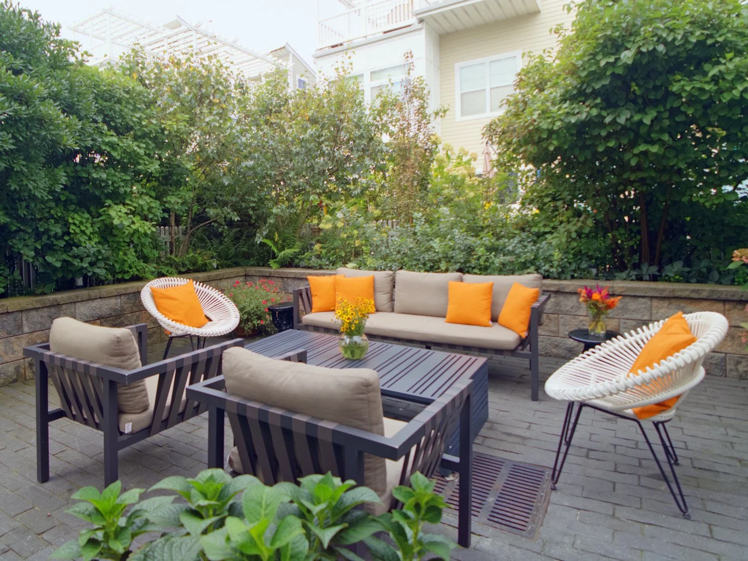 Outdoor patio with beige cushioned patio furniture, chairs with orange pillows, a striped table with a vase of yellow flowers, a small side table with a bouquet, and lush green bushes and trees in the background.