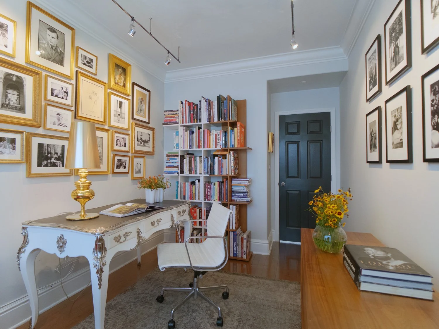 A cozy home office with a white vintage desk, a gold table lamp, a white swivel chair, a bookshelf filled with books, framed photographs on the walls, and a wooden side table with a vase of yellow flowers.