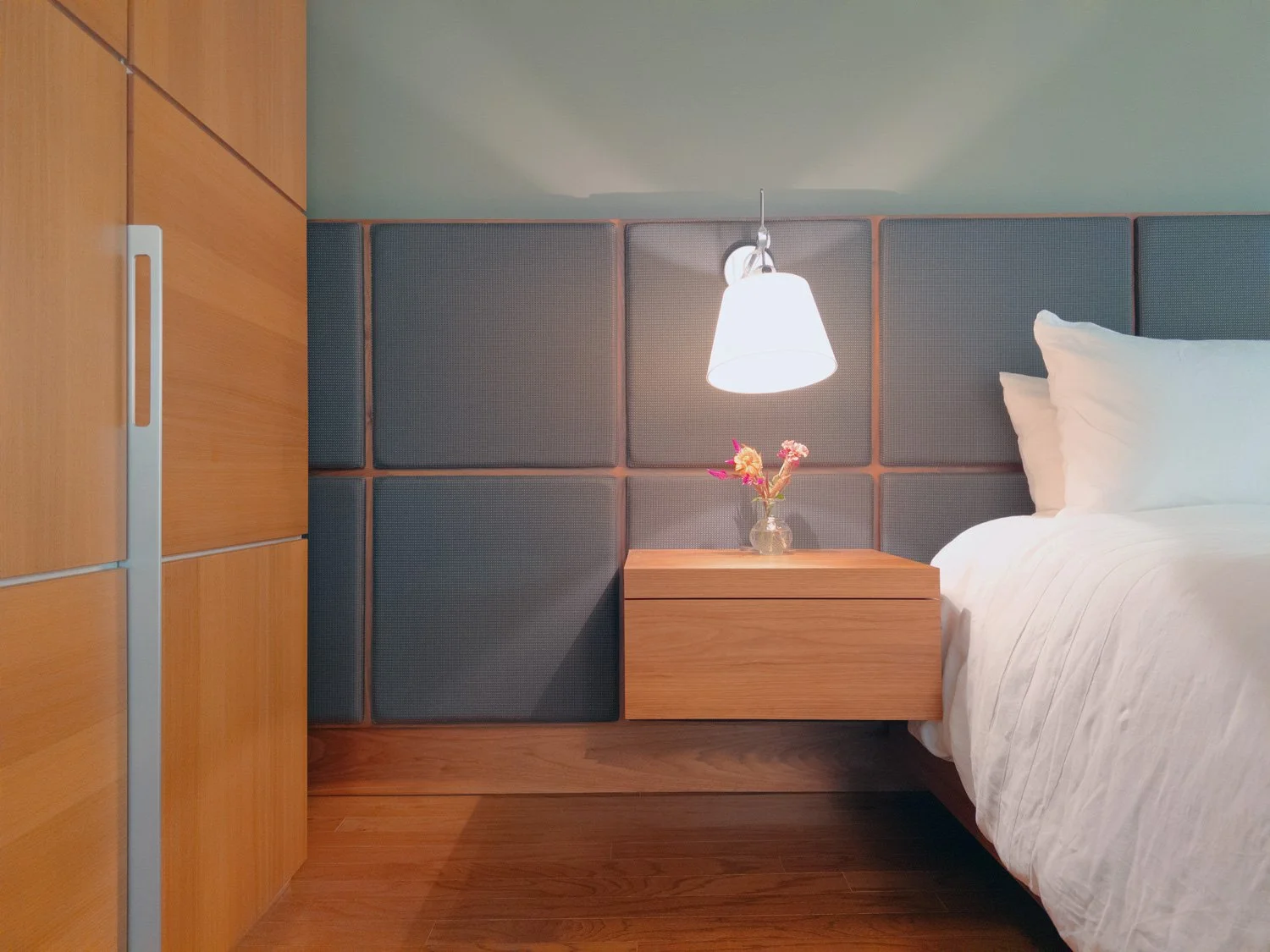 Close-up of a bedroom with a bed, bedside table with flowers, and a wall-mounted reading lamp.