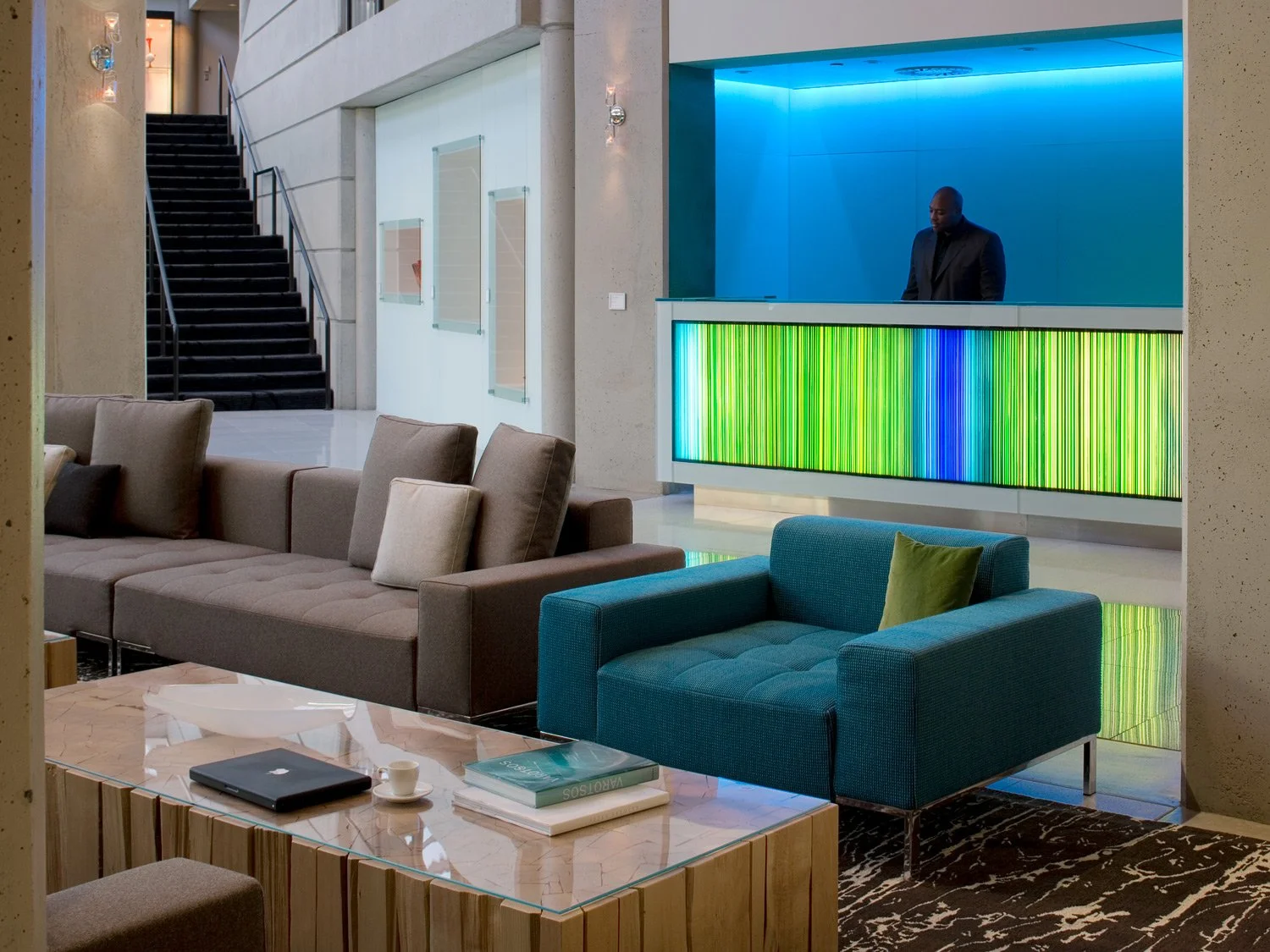 Modern hotel lobby with beige and teal sofas, a marble coffee table, and a colorful digital display on the wall behind a reception desk. There is a man in a black suit standing behind the reception counter, which is illuminated with blue lighting. A staircase with black railings is visible on the left side.