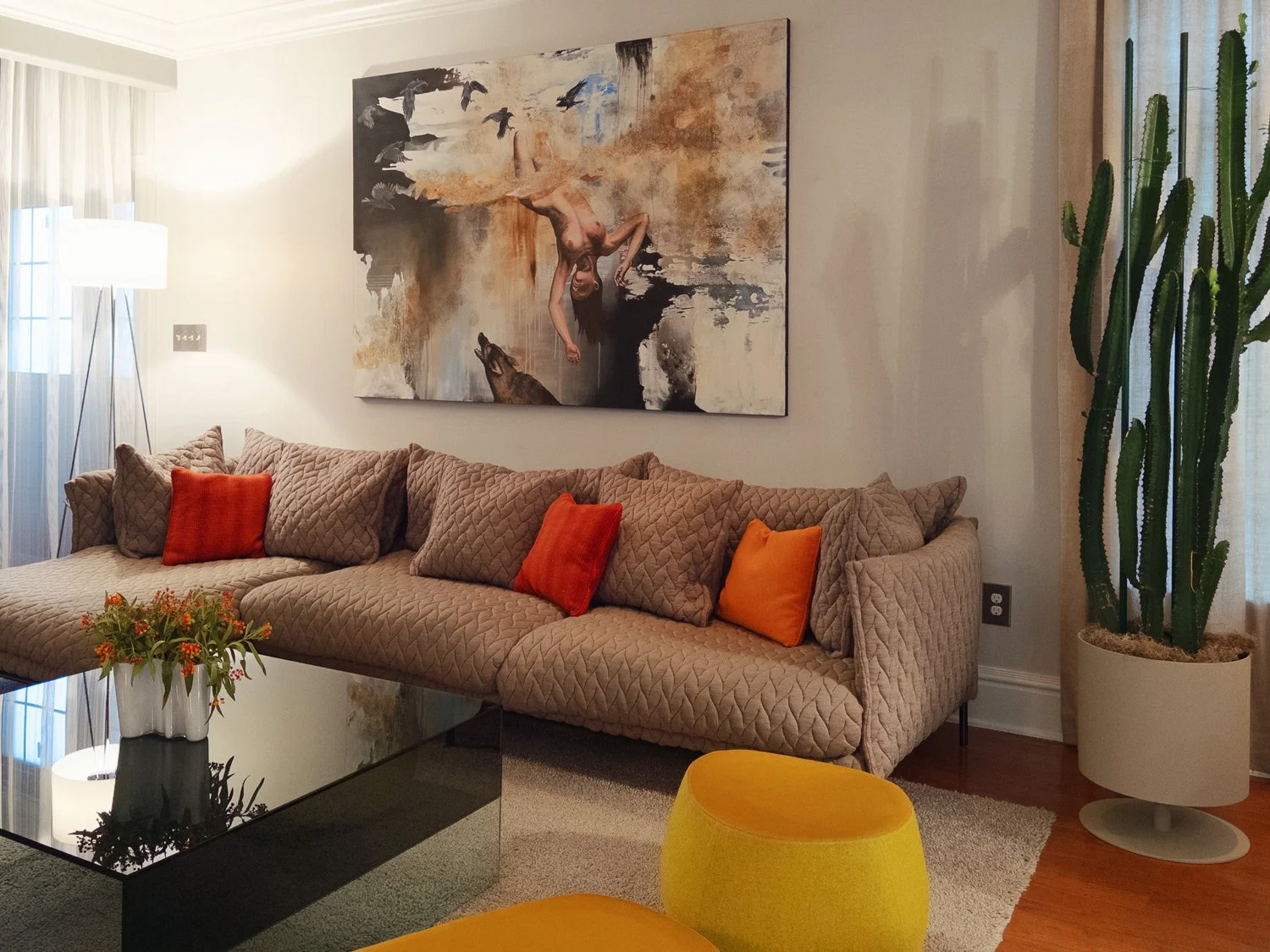 Living room with a beige quilted sofa, three orange accent pillows, a glass coffee table with a flower arrangement, a tall cactus plant in a white pot, a colorful abstract painting on the wall, yellow ottomans, a floor lamp, and sheer curtains in the background.