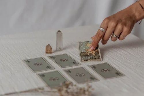 A person with painted nails and rings on their fingers moving a tarot card on a white table, with several other tarot cards placed face down and crystal stones nearby.