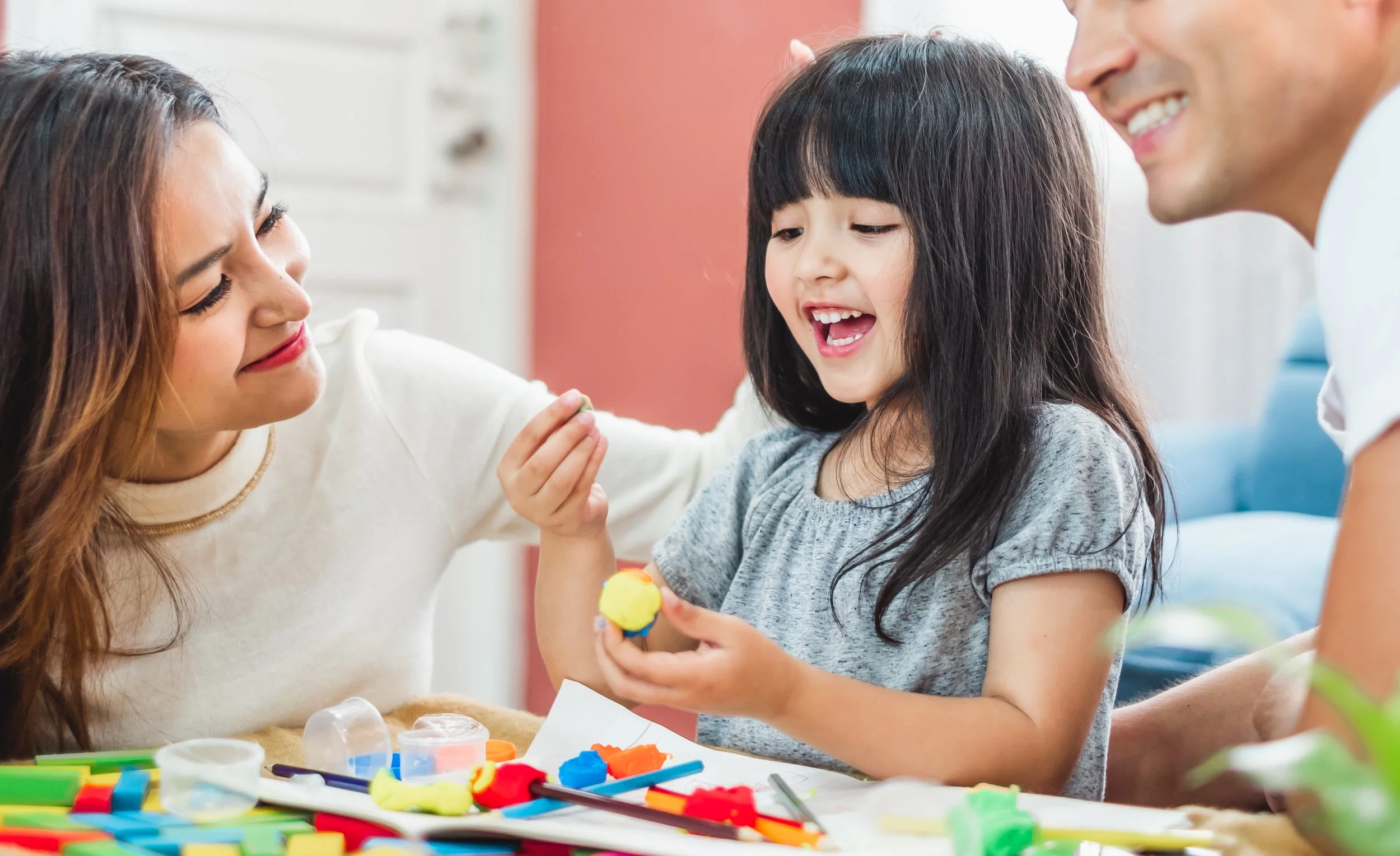 A model  and a young model are sitting at a table with colorful playdough. The girl is smiling and showing her playdough creation to the adults. They seem engaged and happy.