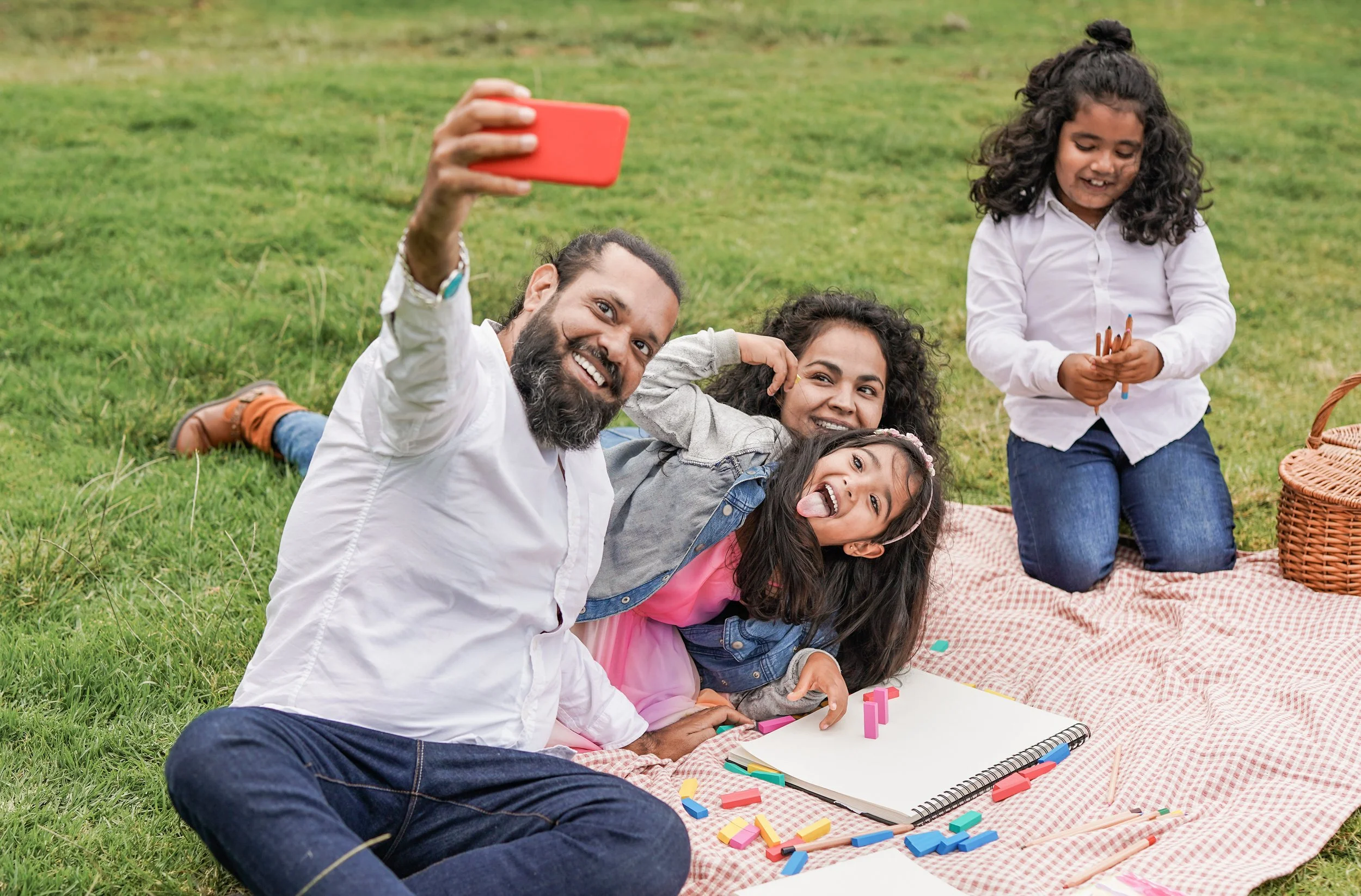 A family smiles while taking a selfie on a picnic blanket.