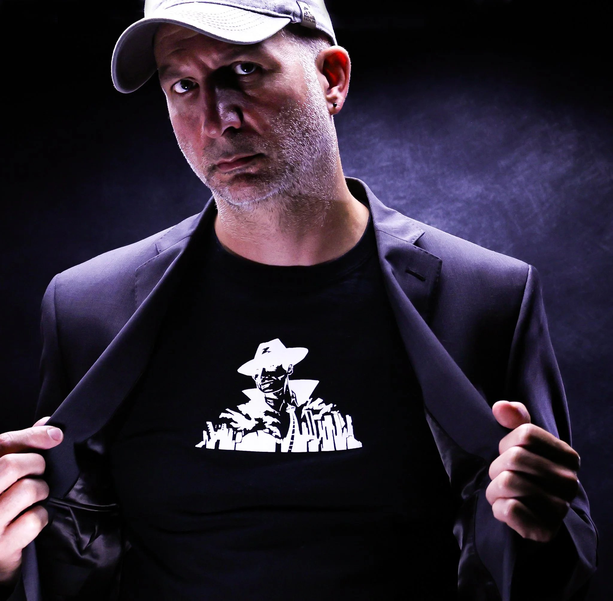 A man with a grey stubble, wearing a light grey cap, black blazer, and black t-shirt with a white graphic of a man in a hat and jacket, revealing the graphic by holding open his blazer.