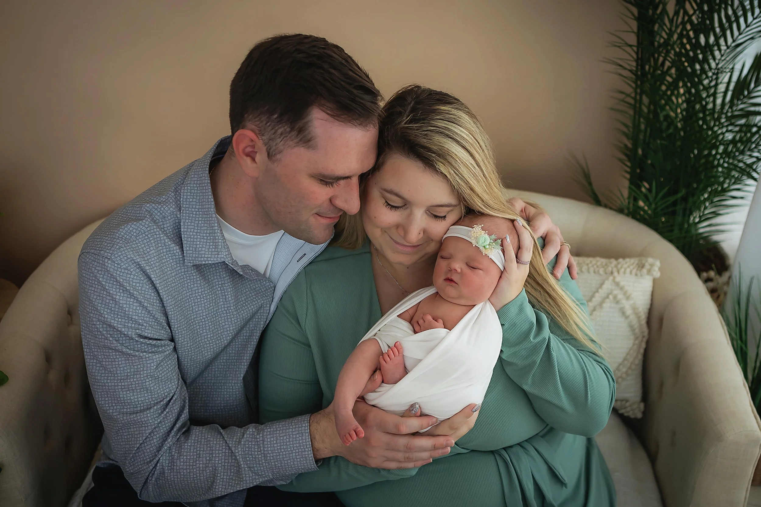 New mom and dad on a neutral colored couch holding their newborn baby girl wrapped in a white wrap with a petite headband with small flowers