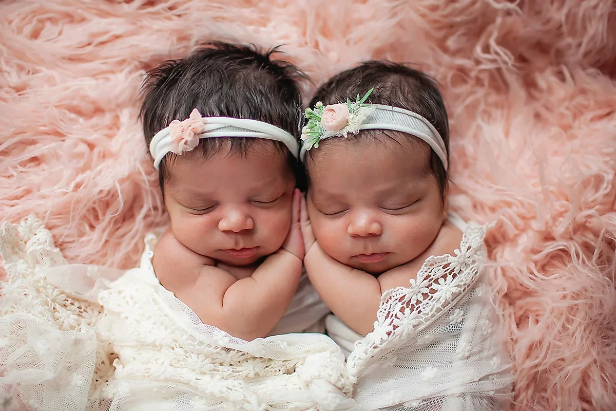 Twin newborn baby girls posed sweetly with heads together and hands snuggled touching between them on a pink fluffy rug with ivory lace wrap.