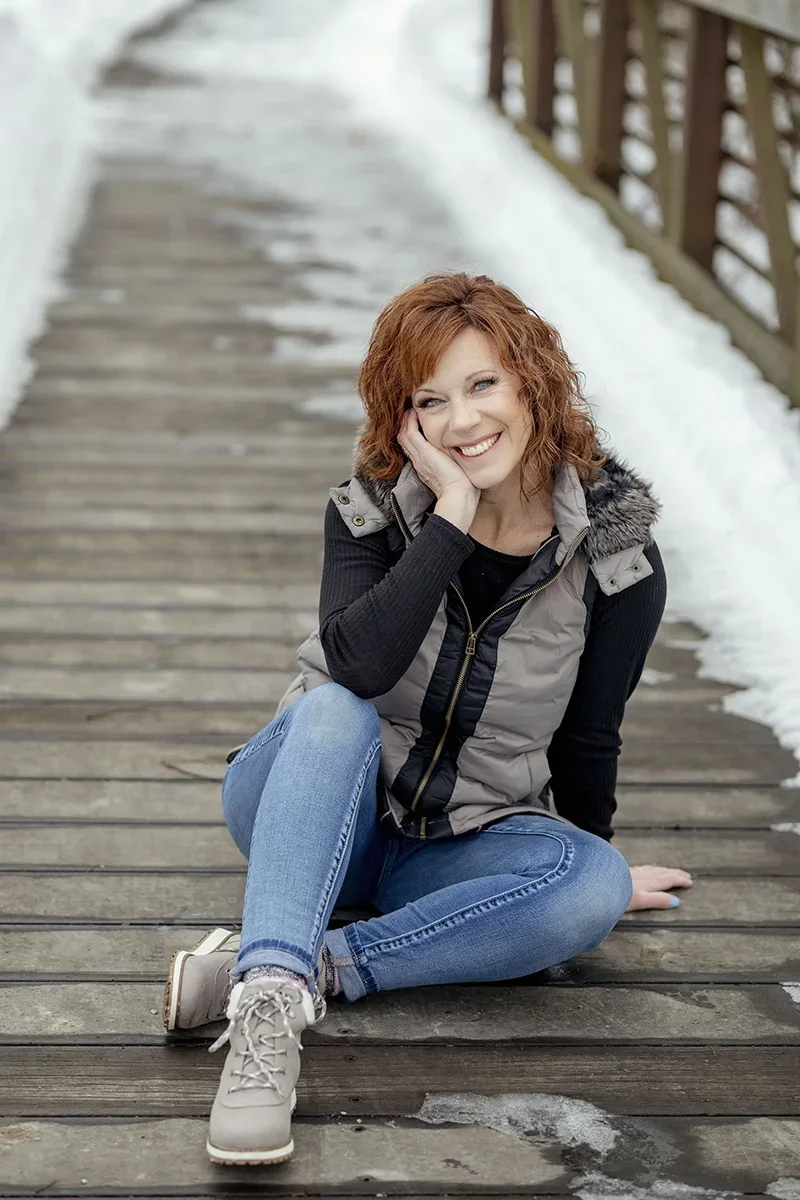 Portrait of Shauna Yenger, owner of Memory Lane Photography, sitting on a wooden bridge outdoors in winter, smiling toward the camera