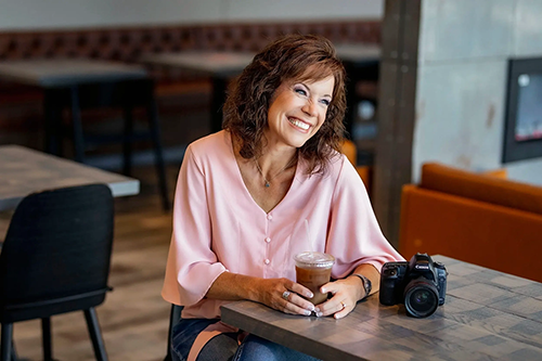 Smiling woman with shoulder-length hair wearing a pink blouse sitting at a table in a cafe, holding a coffee, with a camera on the table.