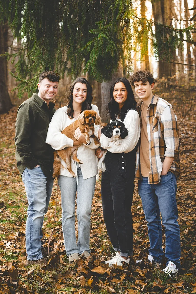 Shauna's children standing outdoors with her family and two dogs during a fall family portrait session in a wooded setting