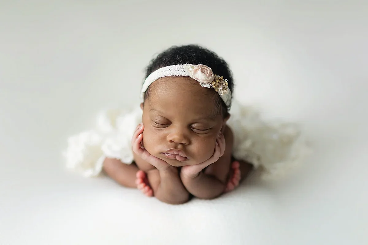 Newborn baby girl with thick, curly hair on white background with lace wrap and floral headband, minimalist newborn photography style.