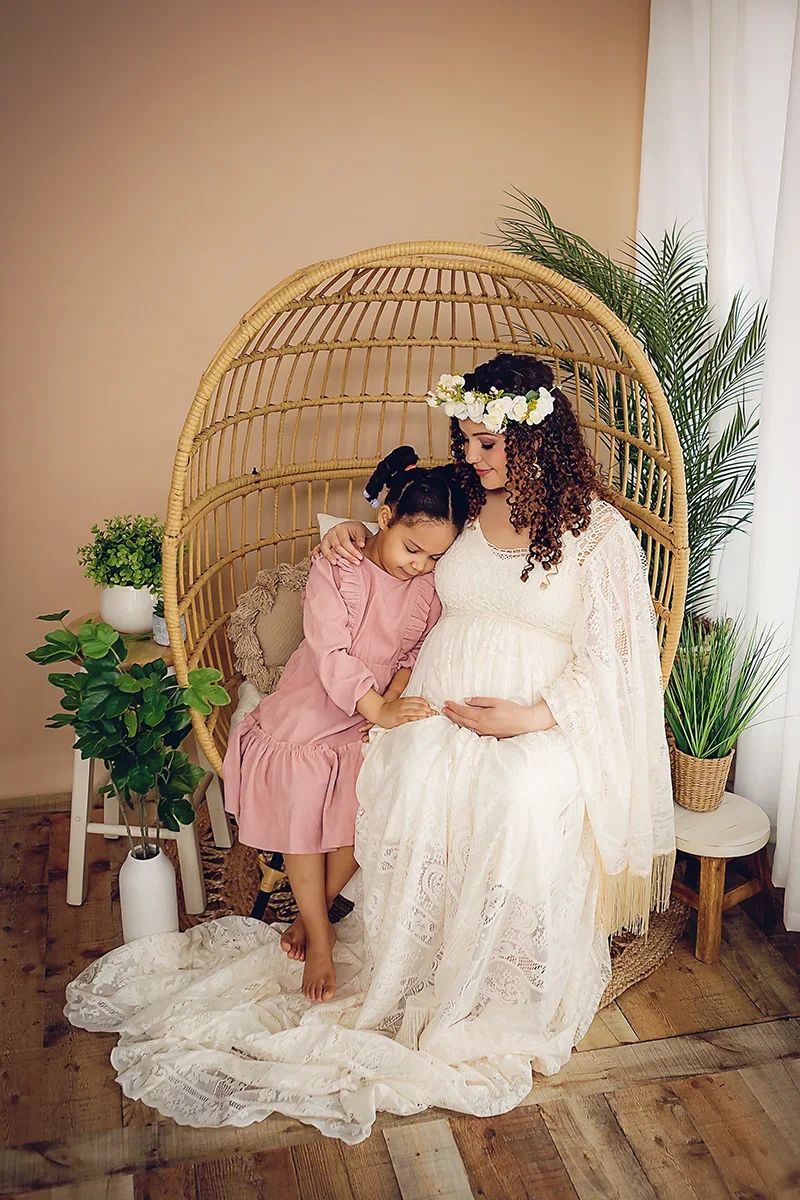 Expecting mother in an ivory maternity gown, sitting in a Boho style chair with her daughter gazing at her expecting belly.
