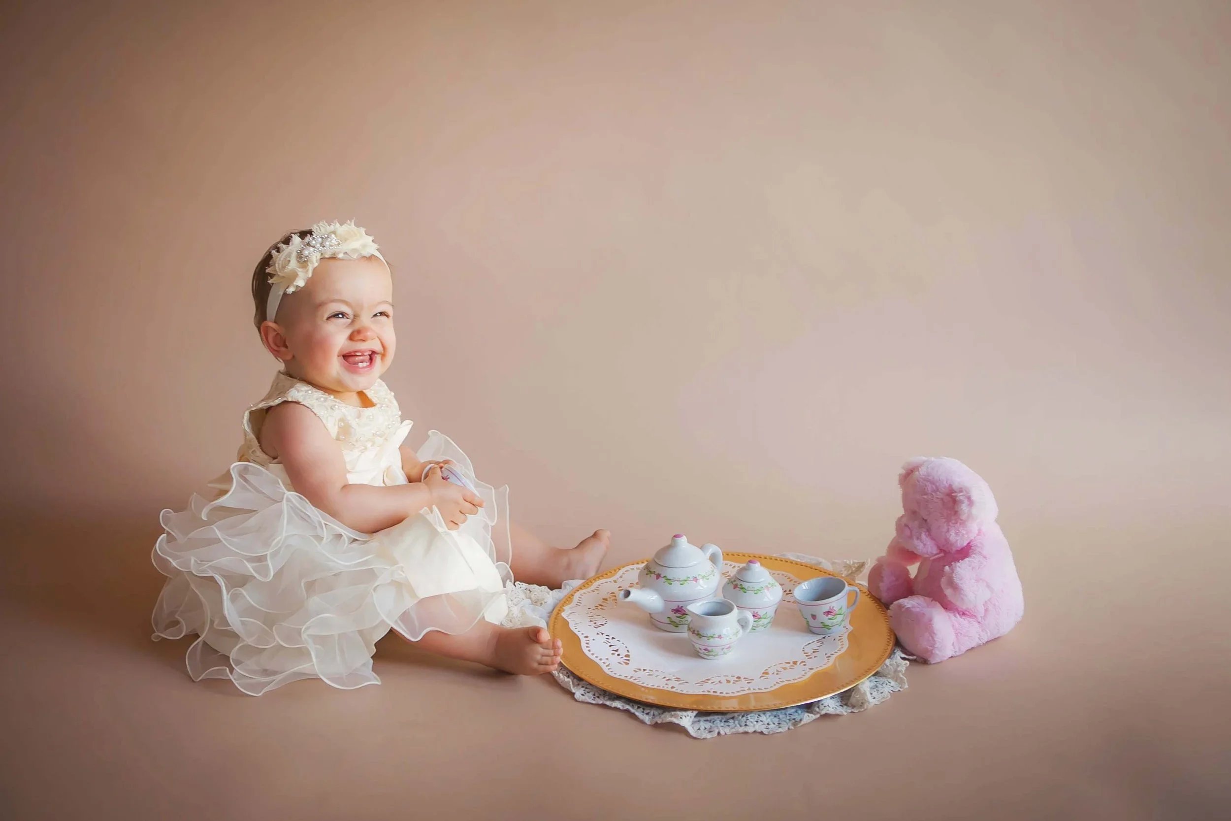 One-year-old baby girl smiling during a milestone portrait session with a tea party setup and soft neutral studio backdrop