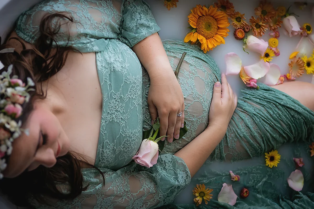 Expecting woman with sage green maternity gown sitting in a luxurious milk bath holding a single pale pink rose wearing a floral crown.
