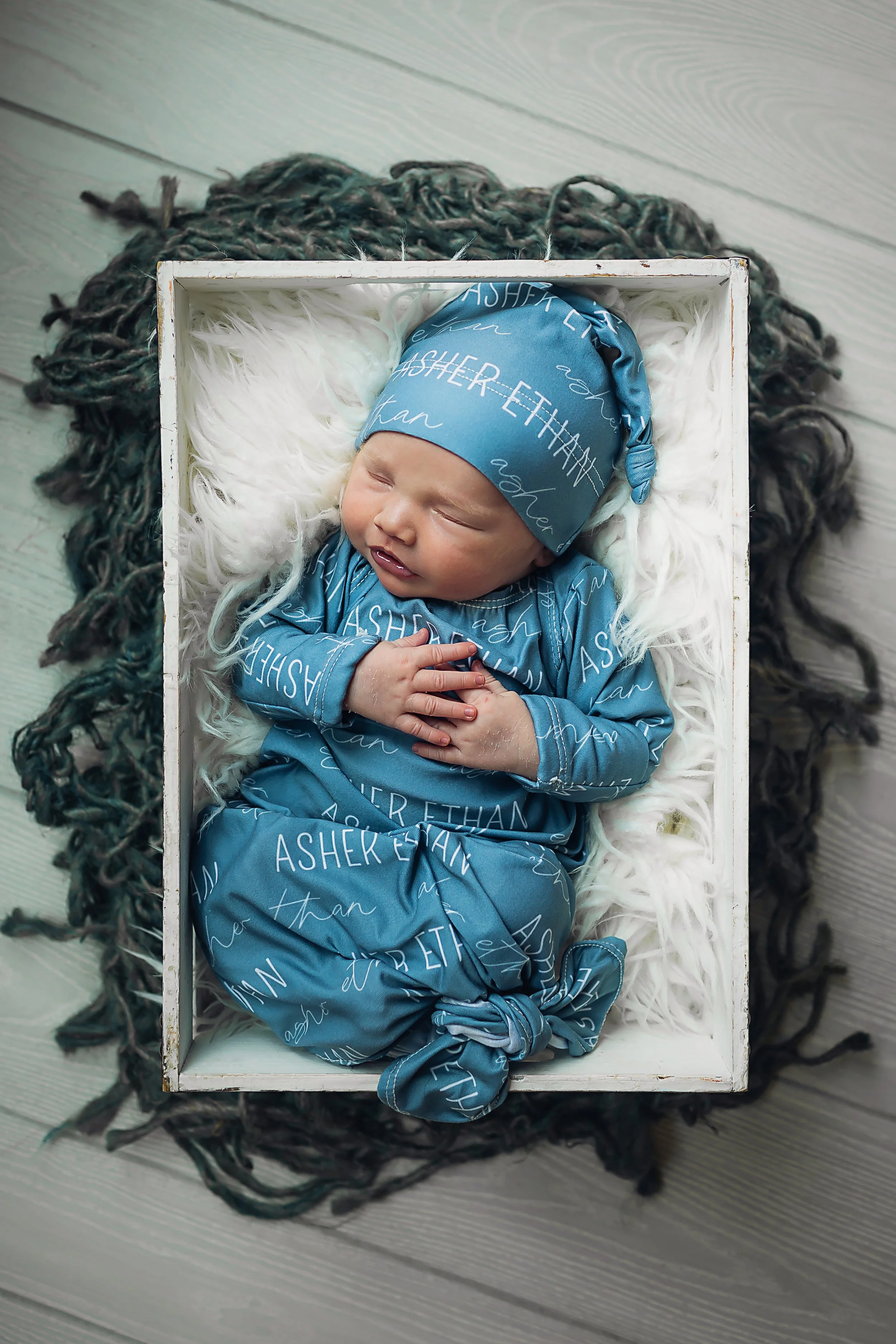 Newborn boy laying in a crate on a white fur layer wearing a custom made sleeper and sleepy hat with his name all over the outfit.