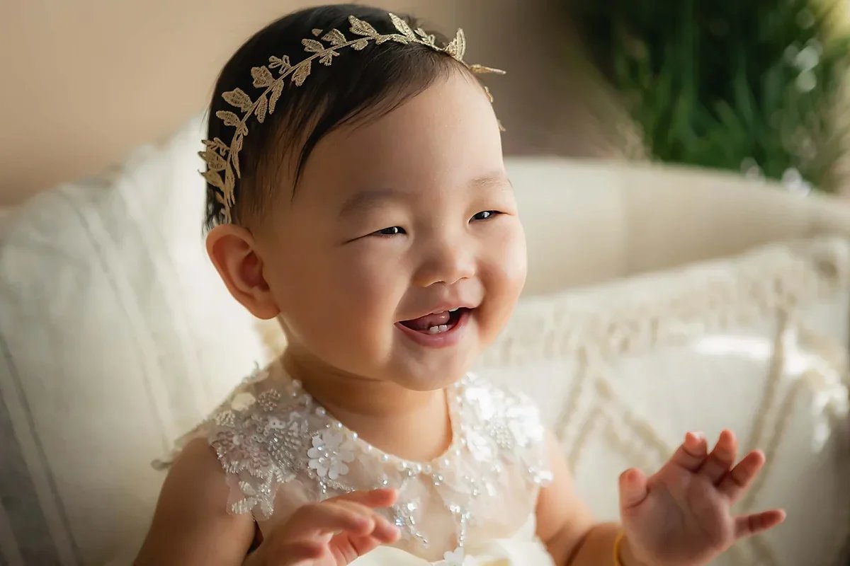 Close-up milestone portrait of a smiling one-year-old baby girl wearing a white dress and floral headband in Indianapolis.