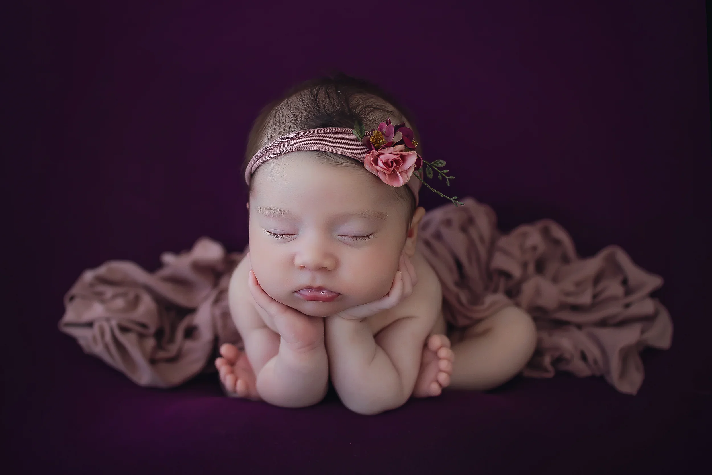 Newborn baby girl nestled snuggly in froggy pose on a burgundy background with a pink floral headband.
