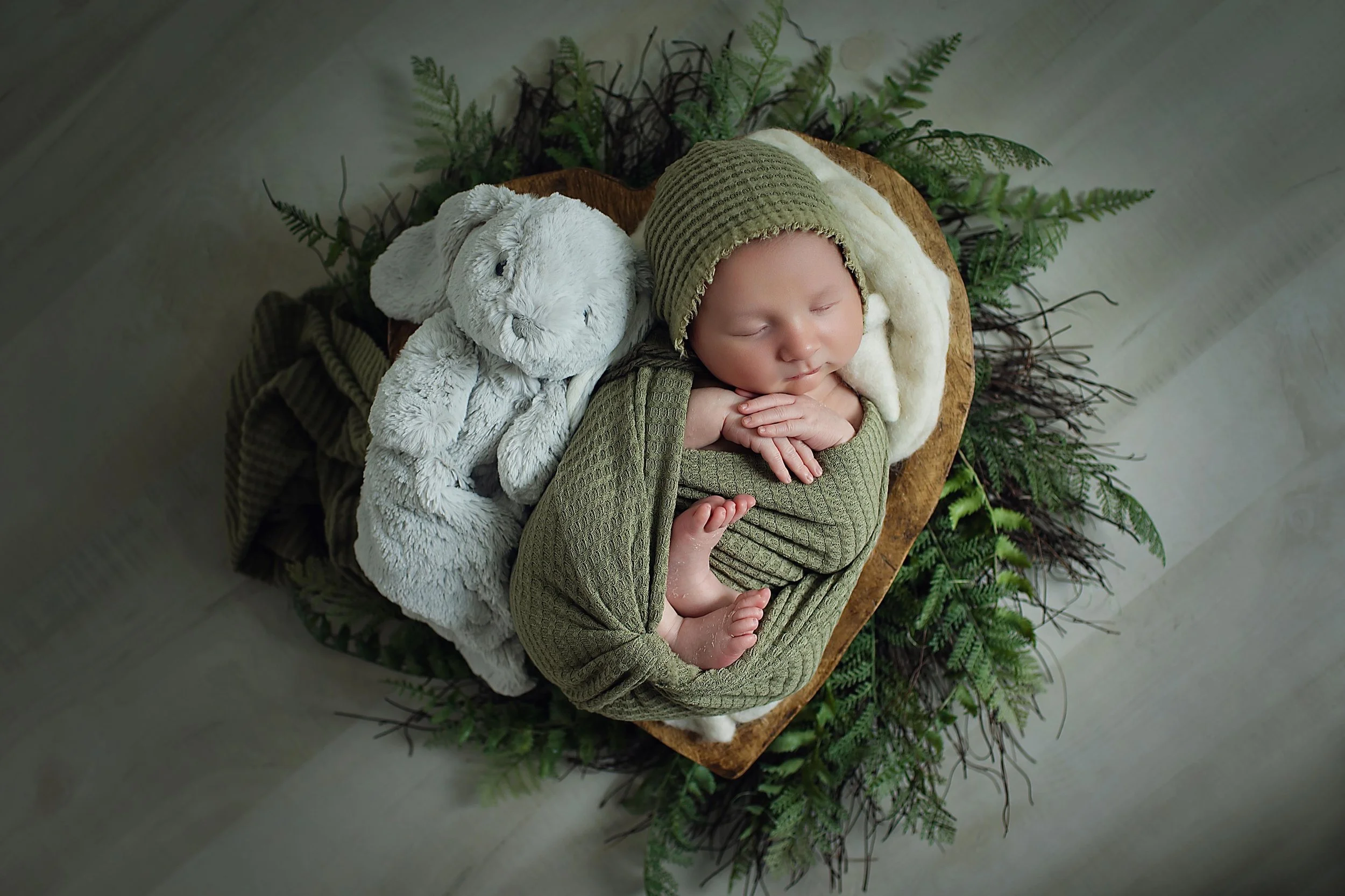 Newborn Baby boy posed in olive green on greenery with a plush bunny snuggle up at his side
