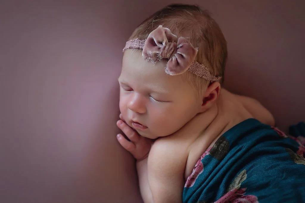 Indianapolis newborn photo session showing baby girl wearing a mauve bow covered in a teal floral wrap laying on a mauve background.