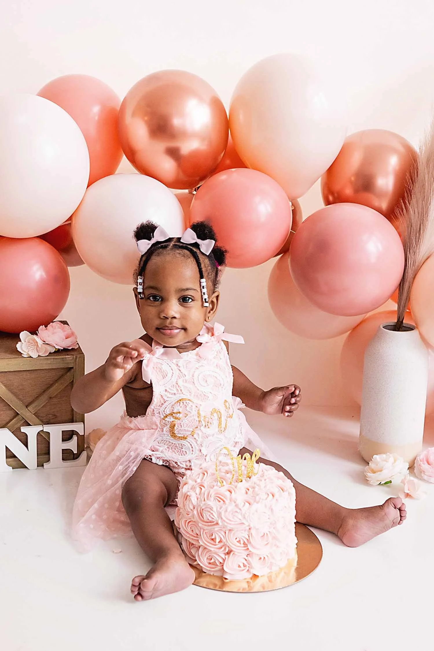 One year old little girl in pink dress sits in front of a balloon arch with a beautiful pink rosette birthday smash cake.