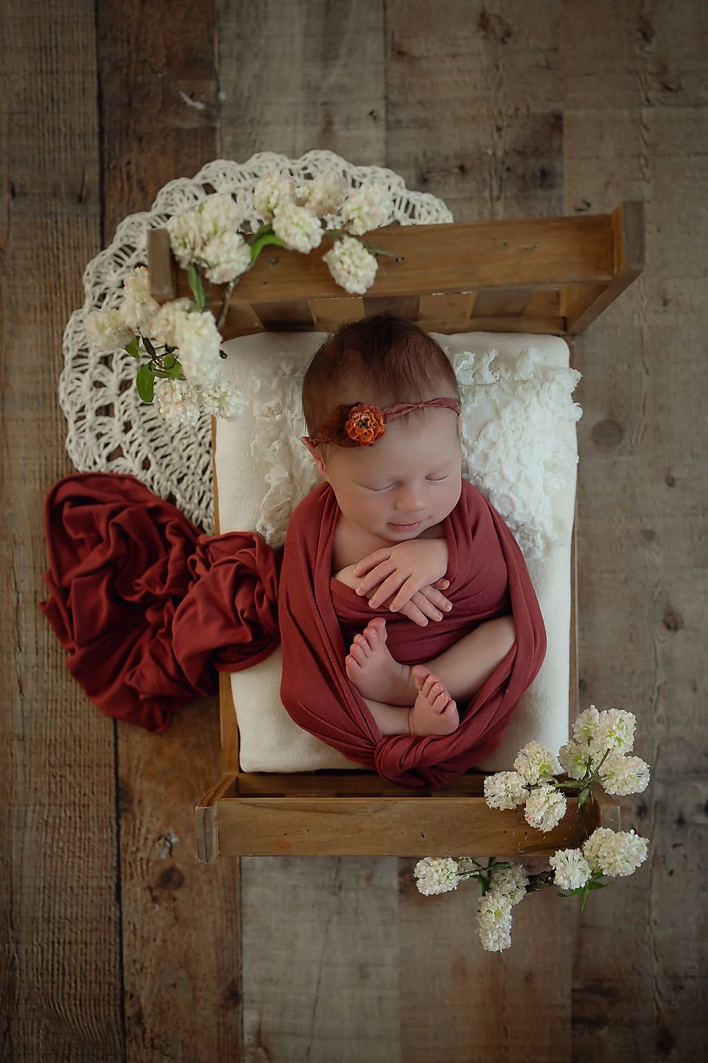 Newborn baby girl swaddled in rust colored wrap laying on a bed with flowers surrounding her sitting on a barn wood floor during a newborn session in Indianapolis, IN.