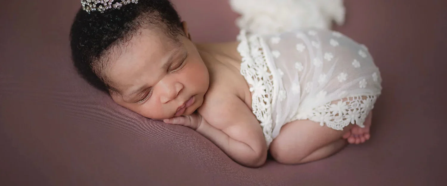Newborn girl sleeping curled up and wrapped in lace ivory fabric while sleeping on rose colored blanket.