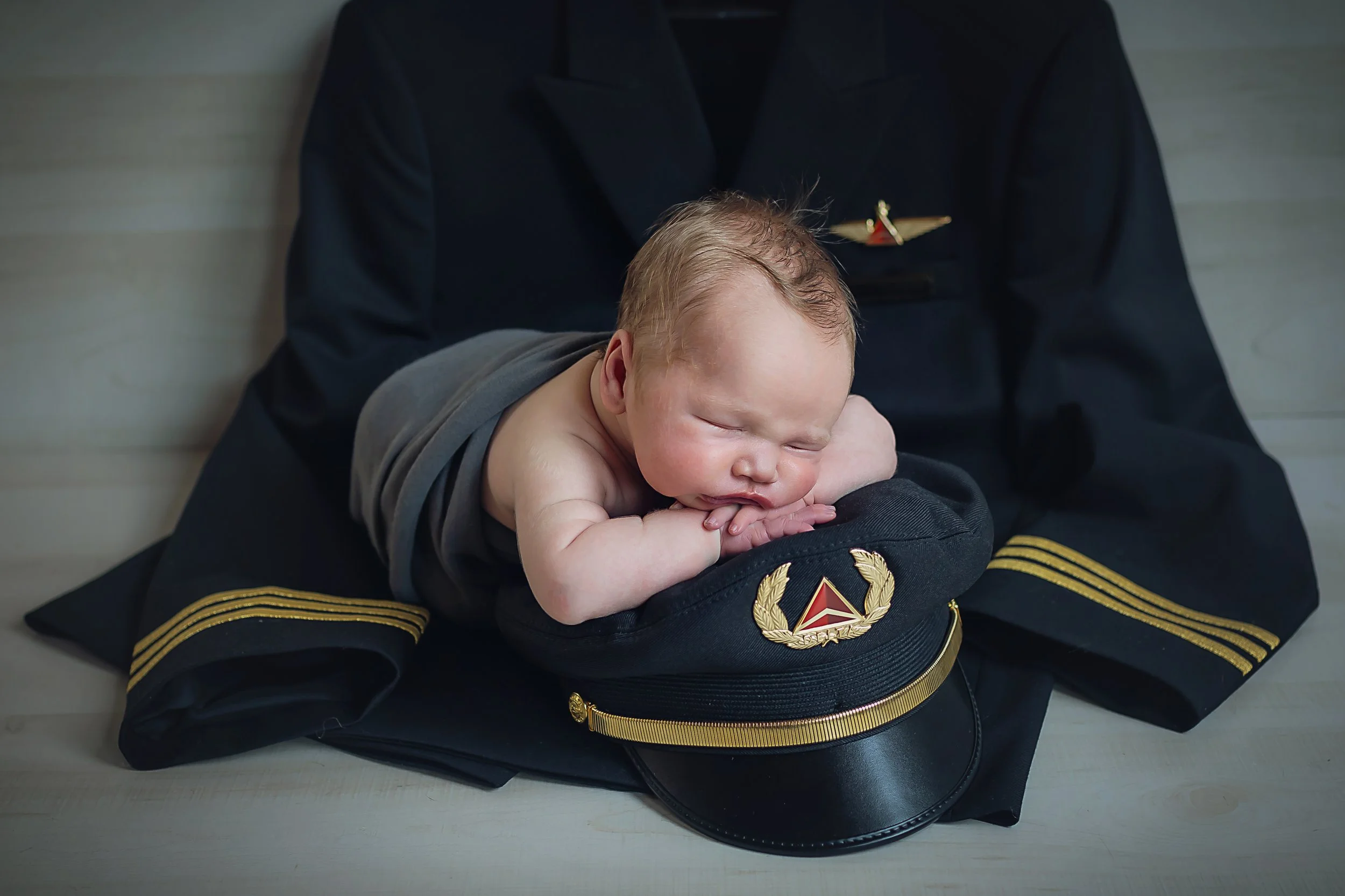 Newborn baby boy laying across his father's pilot uniform with his head resting on his arms on top of dad's pilot hat.