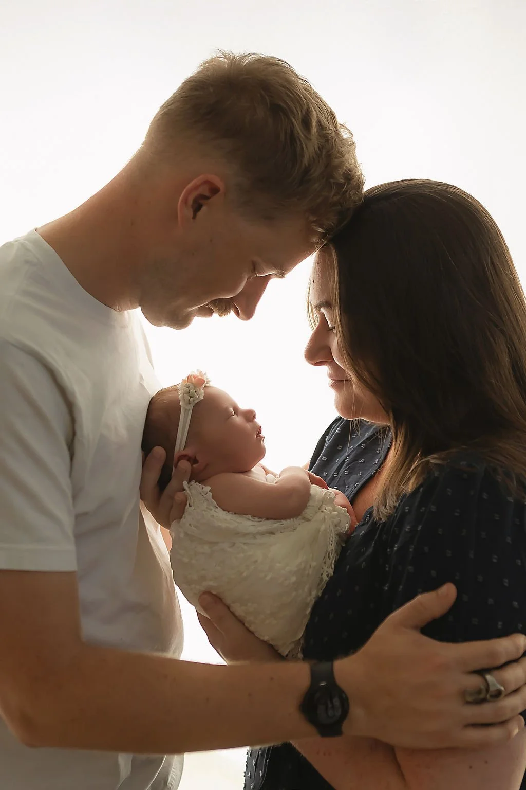 Father and mother touching heads while mom holds newborn baby girl between them at their newborn photography session in Indianapolis.