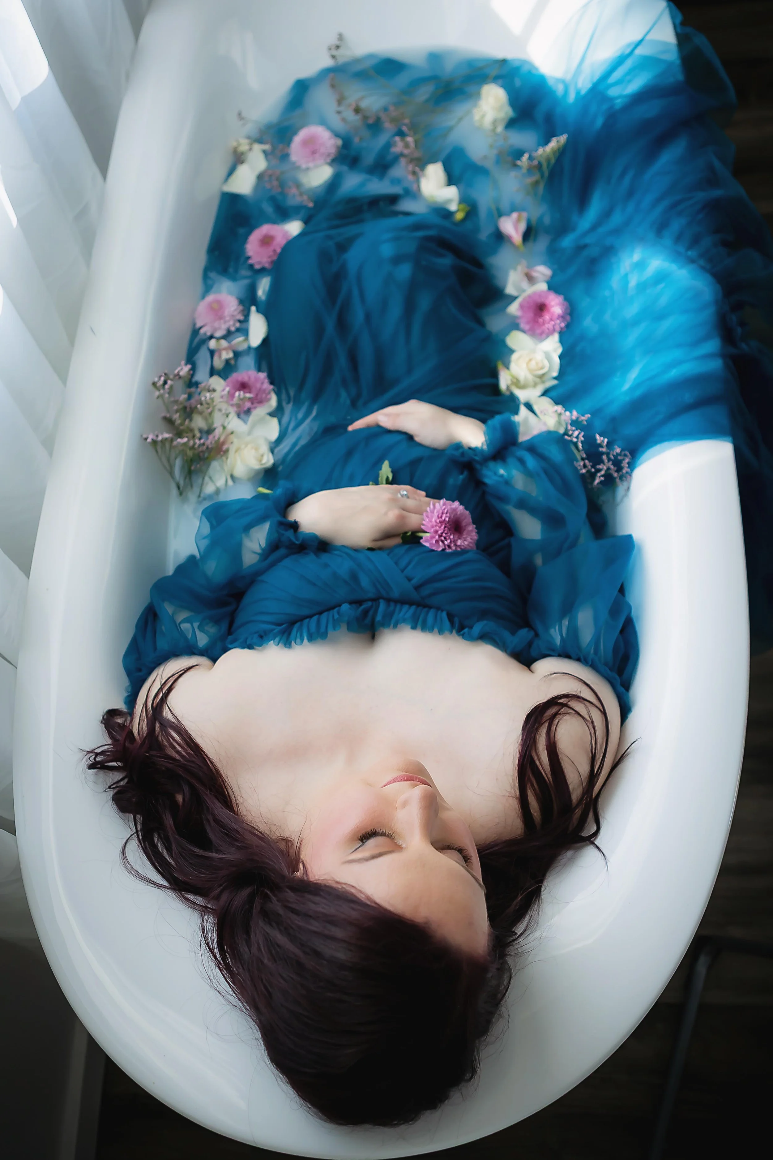 A woman in a blue dress is lying in a white bathtub filled with flowers.