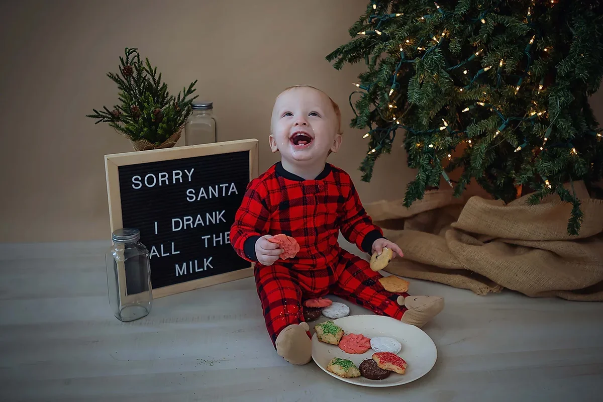 Toddler sitting near a Christmas tree during a holiday milestone portrait session, wearing red pajamas and holding cookies in a studio setting