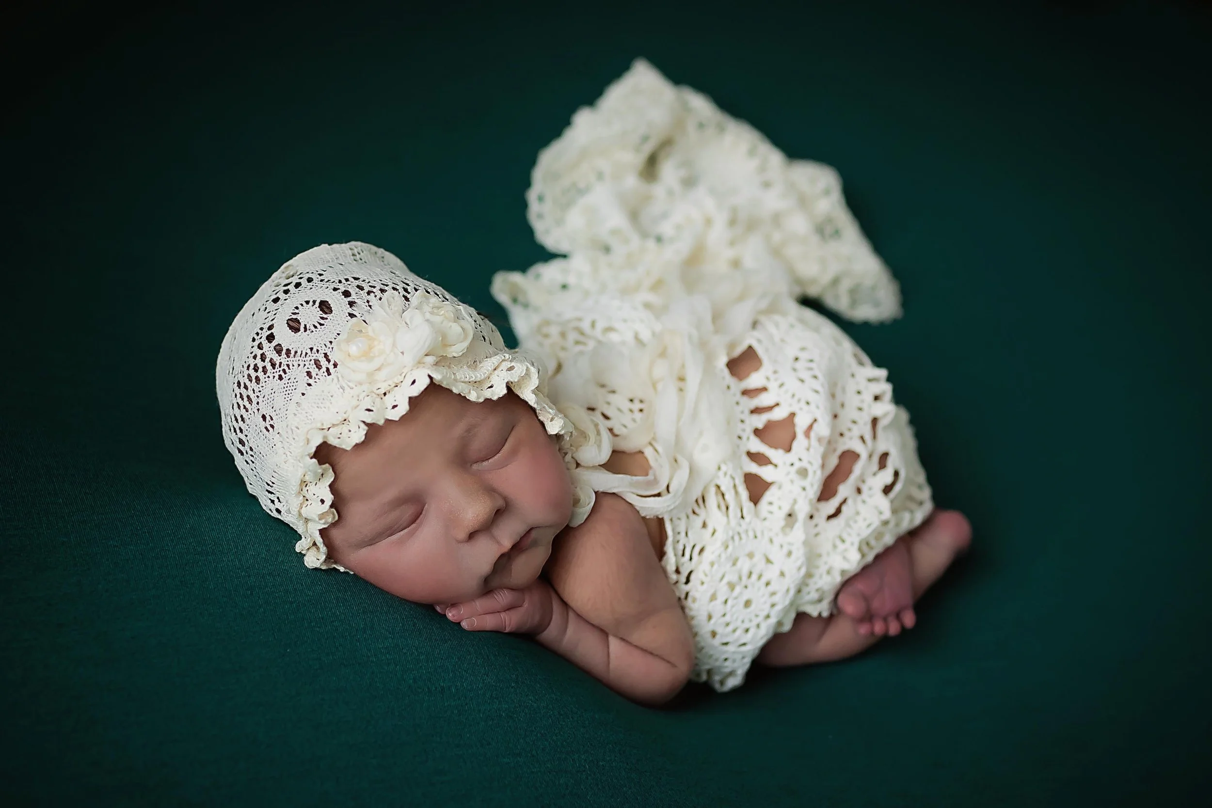 Newborn baby girl wrapped in cream, vintage lace  and  bonnet posed on forest green backdrop in Indianapolis studio