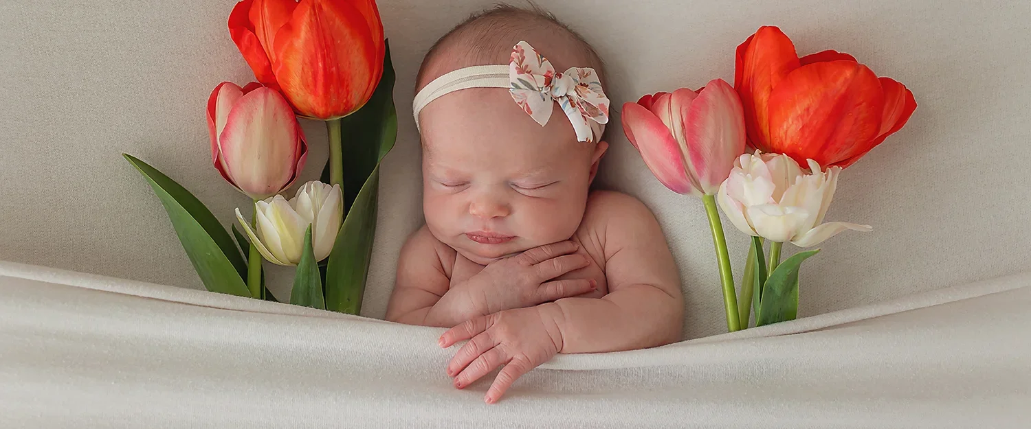Newborn baby girl with bow headband laying tucked in with colorful tulips on either side of her.