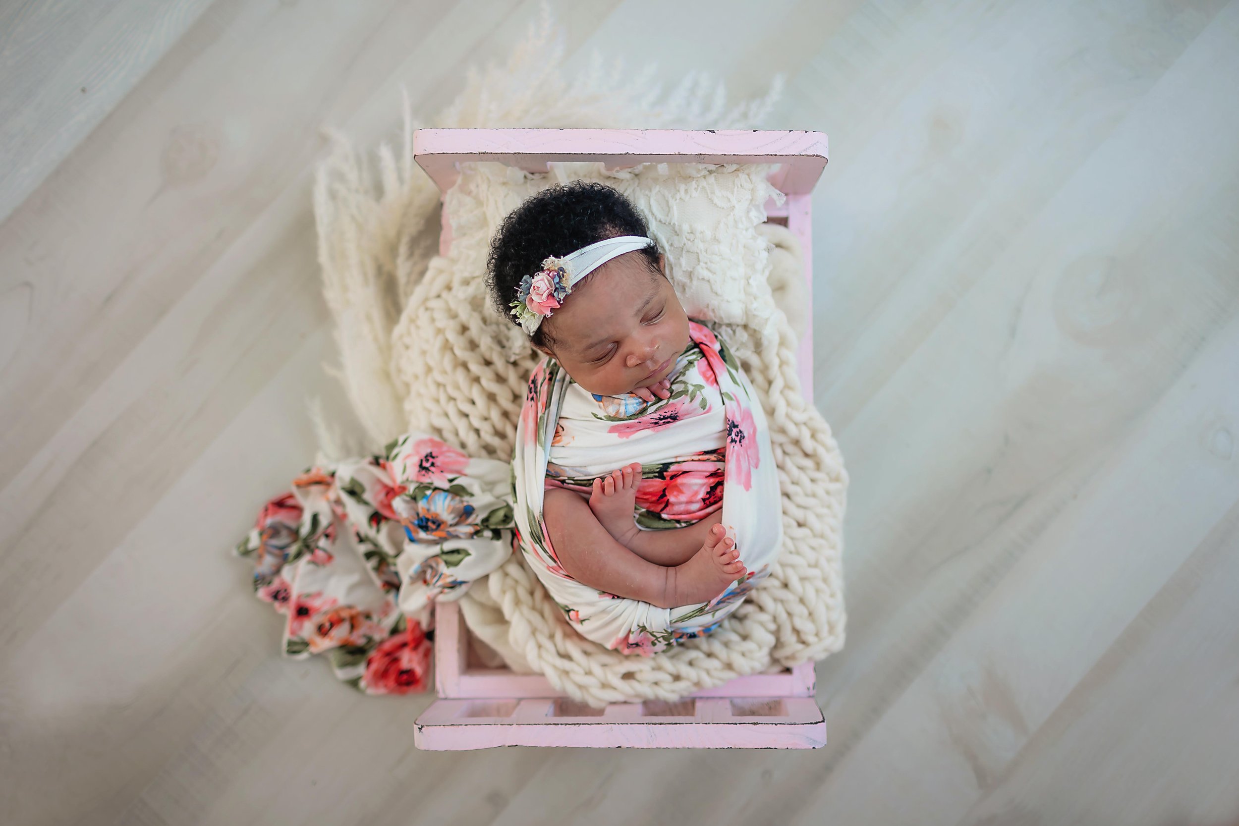 Newborn baby girl posed in a little soft pink bed with cream layers and wrapped in a spring floral wrap with a matching headband