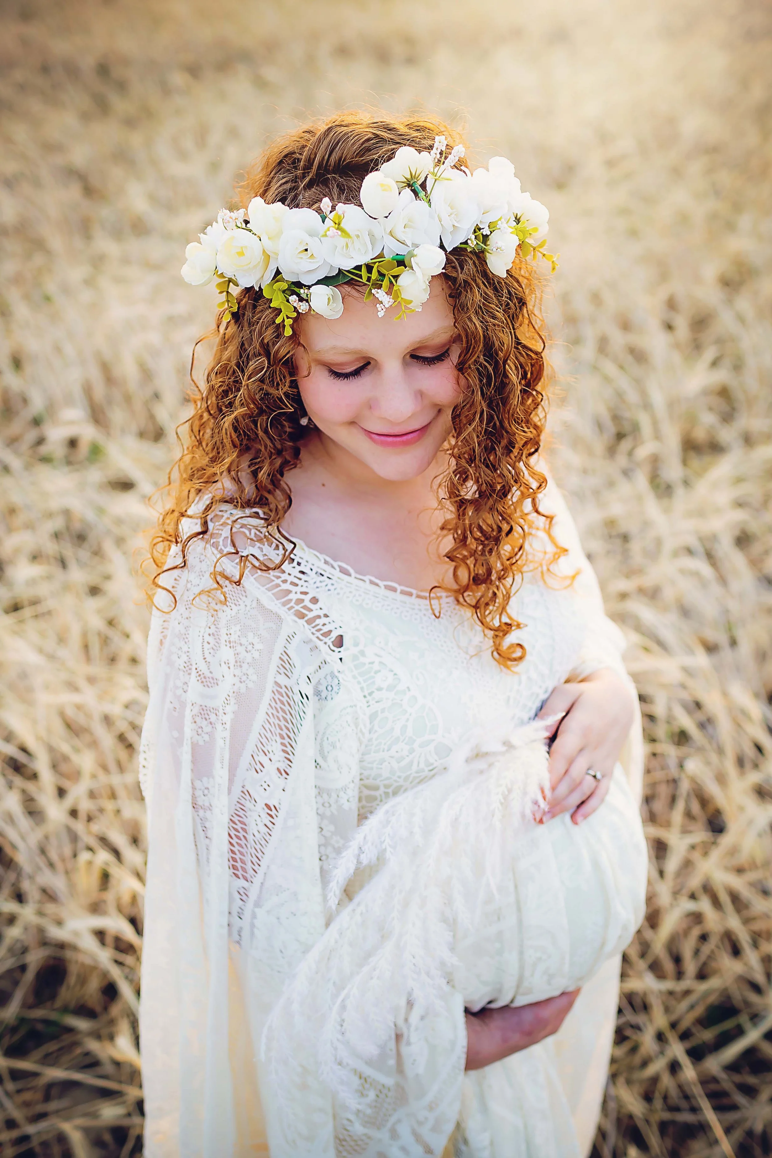 Expecting mother with long curly red hair wearing a flower crown with cream flowers and a boho cream lacy dress holding dried pompass  grasses in her hand  in a field of dry grasses at sunset on the east side of Indianapolis